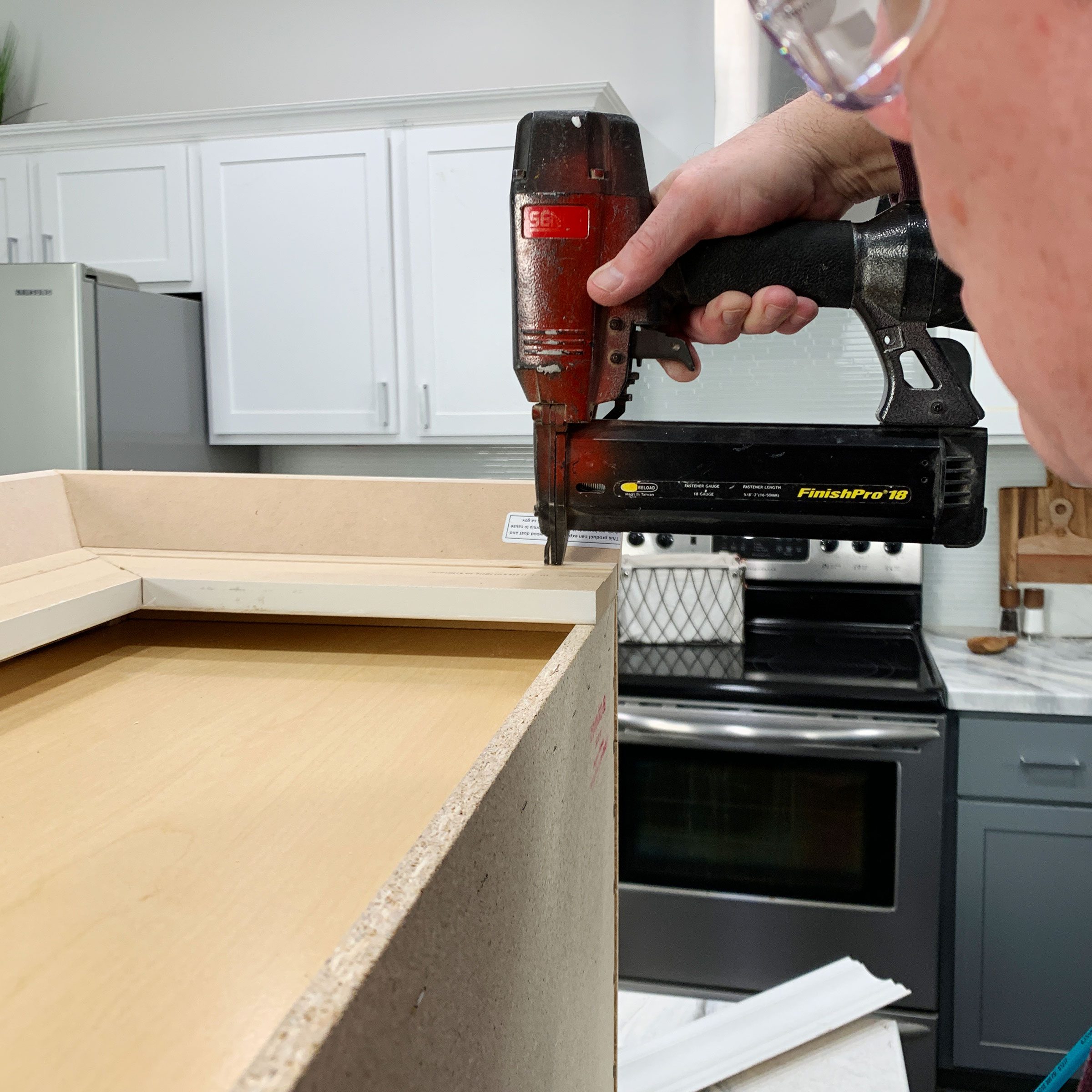 A hand holds a nail gun, securing a board to a wooden structure in a kitchen with metal appliances and a stove in the background.