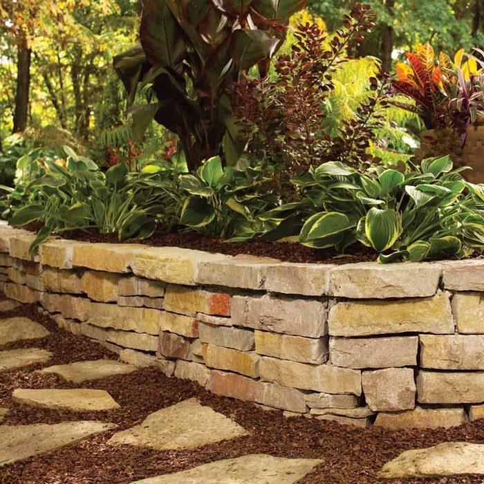 A stone wall borders a garden bed filled with vibrant plants, while irregular stone pathways are laid amidst dark mulch, surrounded by lush greenery.