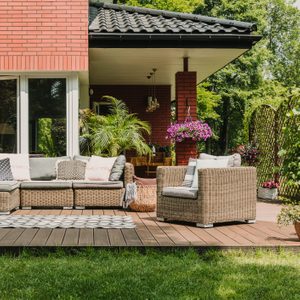 A cozy outdoor seating area features a woven sectional sofa and chair on a wooden deck, surrounded by greenery and potted flowers under a sunny sky.