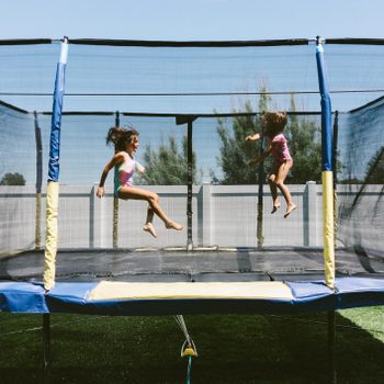 Two girls are mid-jump on a trampoline, surrounded by a grassy area and a white fence. A clear blue sky overhead enhances the playful atmosphere.