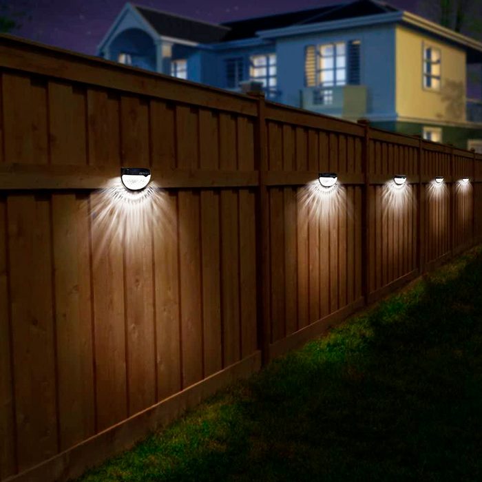 A row of solar lights is illuminating a wooden fence at night, casting beams of light onto the grass, with a house visible in the background.