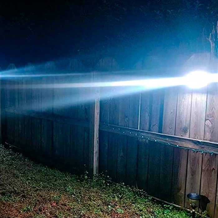 A bright beam of light illuminates a wooden fence at night, creating a striking contrast with the dark surroundings and outdoor grass.