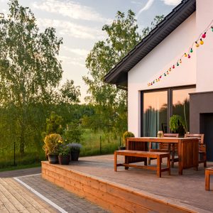 A wooden deck features a table and chairs, surrounded by potted plants, with colorful lights strung above, set against a lush green landscape.