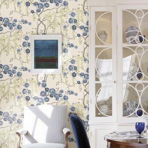 A patterned armchair sits in a sunlit room, with floral wallpaper and a modern art piece framed on the wall beside a glass cabinet.
