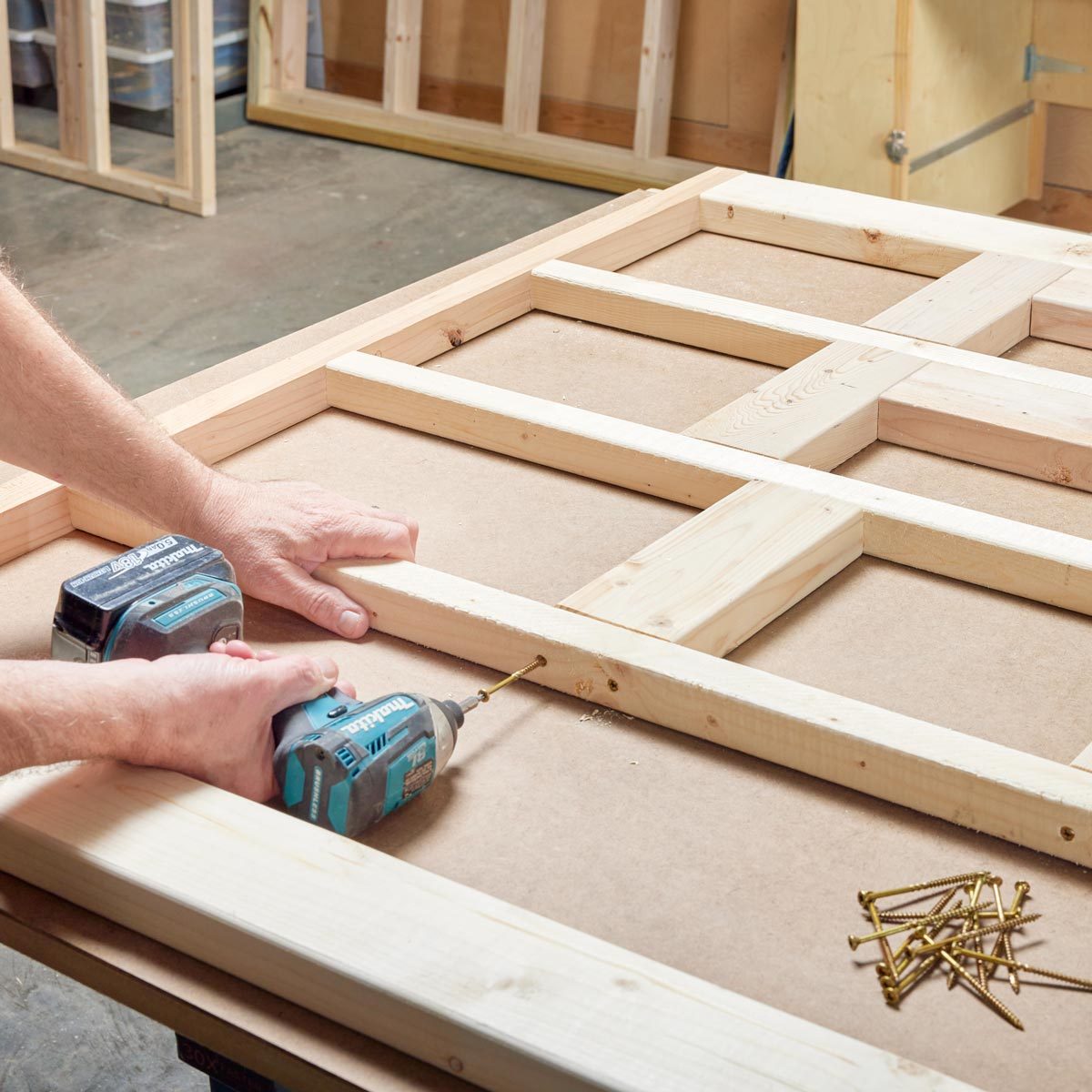 Hands use a drill to secure wooden frames onto a flat surface, surrounded by a workshop environment with tools and wooden panels.