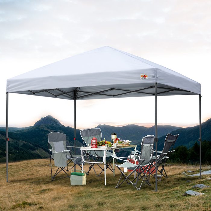 A white canopy stands over a table set with food and drinks, surrounded by folding chairs on a grassy hill, with mountains in the background.