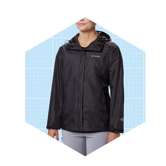A person wears a black rain jacket, smiling, against a light blue grid background. The jacket features a hood and a logo on the chest.