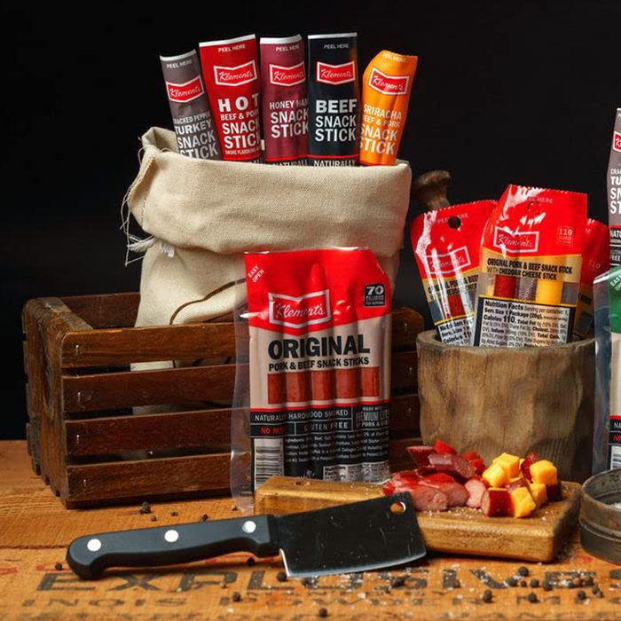 Snack sticks are displayed in various packages, while a cutting board holds sliced meats and cheese. A wooden crate and knife are nearby on a rustic surface.