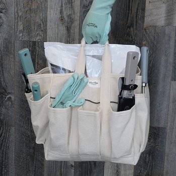 A canvas gardening tote holds various tools, including gloves and garden items, while a hand reaches to grasp one of its handles. The background features wooden flooring.