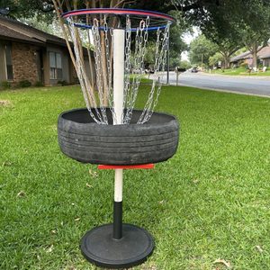 A disc golf basket constructed from a tire and chains stands on a grass lawn, surrounded by residential houses and a quiet street in the background.