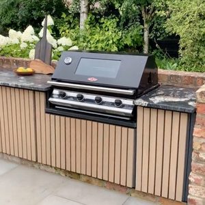 A black grill sits on a granite countertop surrounded by wooden cabinetry, with a bowl of fruit nearby, all set in a green garden environment.