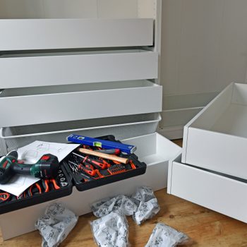 A disassembled white drawer unit sits on wooden flooring, tools and assembly instructions scattered nearby, while two drawers are partially pulled out.