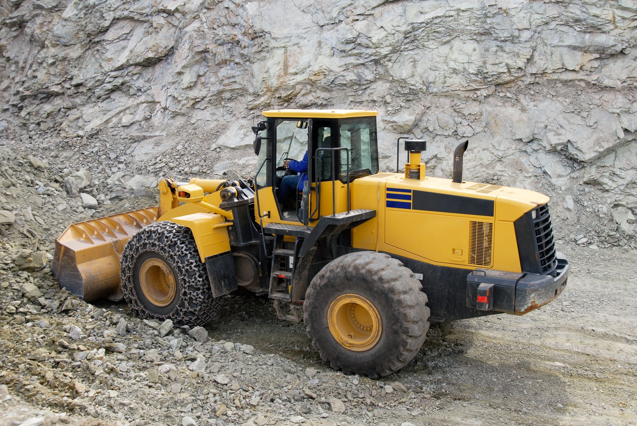 A yellow bulldozer operates in a rocky quarry, moving earth while a driver navigates from the enclosed cabin, surrounded by uneven, gravel-strewn terrain.