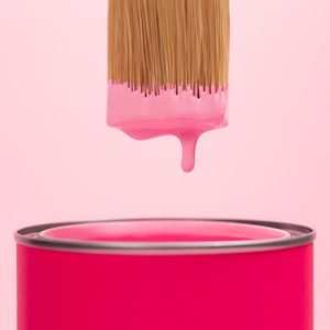 Pink paint is dripping from paintbrush bristle into can with pigment against pastel pink background