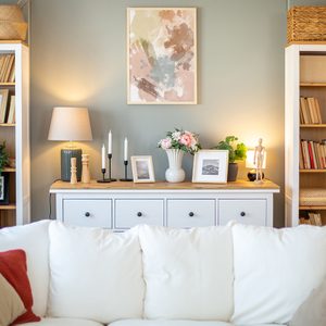 Living room with white cozy sofa, white-wooden chest of drawers with many decoration pieces between two bookshelves with books and wicker baskets against gray wall.