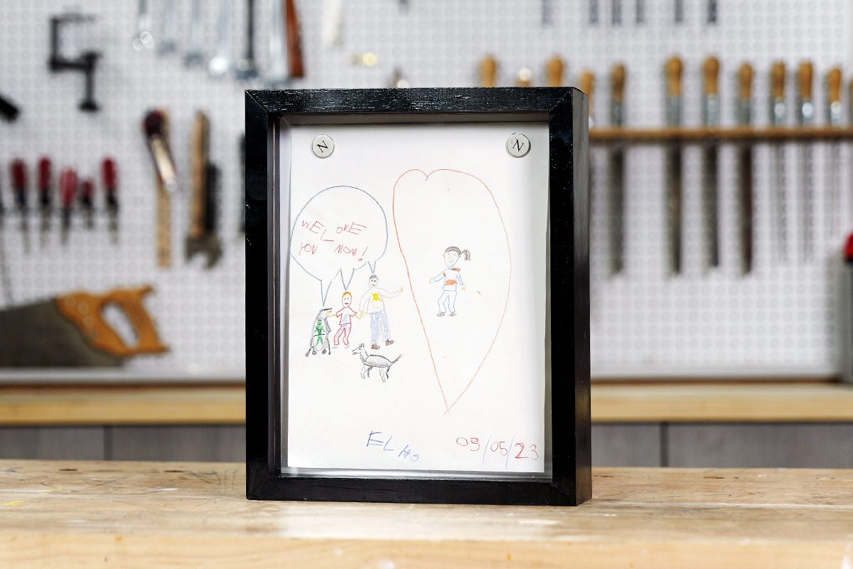 Framed child’s drawing showing two large hearts with small figures inside, sitting on a wooden table. The background features a workshop wall with hanging tools like saws, screwdrivers, and hammers.
