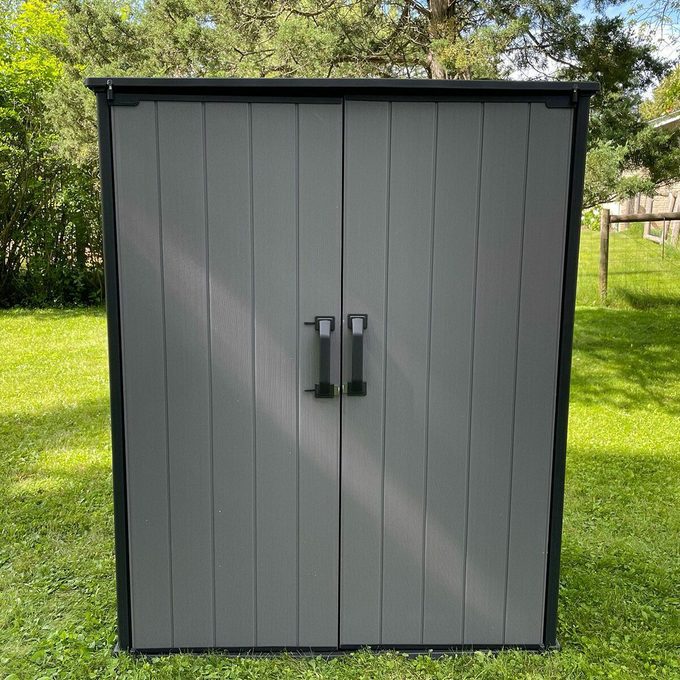 A gray outdoor storage shed with double doors and two black handles, standing on green grass with trees and a fence in the background.