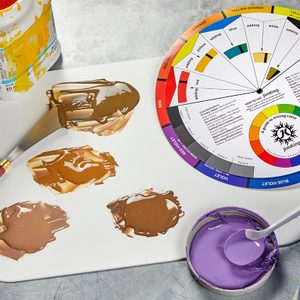 A paint mixing palette with brown and tan shades, a palette knife, jars of blue, yellow, and purple paint, a white mixing tray, and a color wheel displayed on a metallic surface.